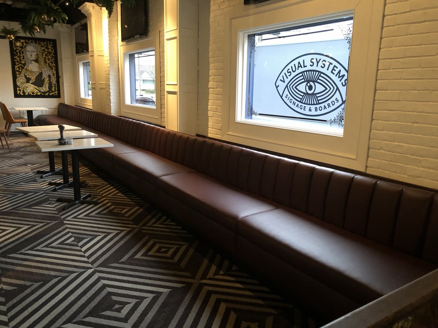 Long brown channel-tufted banquette bench running along a brick wall above striped carpet