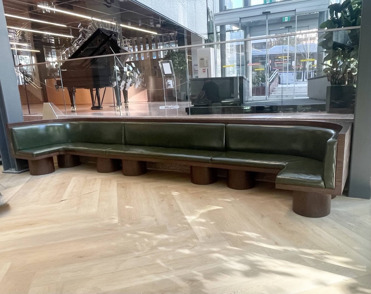 Olive green leather bench on round wood plinths in a bright lobby overlooking a grand piano